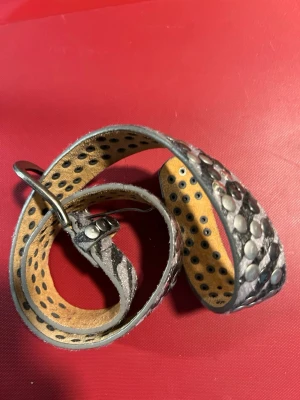 Silvrigt nitskärp med prickigt mönster - Säljer ett coolt bälte i silverfärgat skinn med nitar och prickigt mönster. Bältet har metallspänne och detaljer som ger en edgy look. Perfekt för att lyfta vilken outfit som helst.