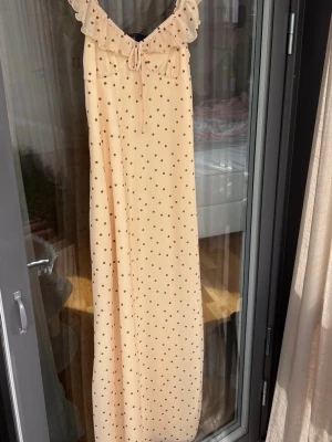 Ljusgul prickig långklänning med volang - Supersöt långklänning i ljusgul ton med små svarta prickar. Klänningen har volangdetalj vid urringningen, knyt framtill och smala axelband. Materialet känns lätt och flowy, perfekt för varma dagar.