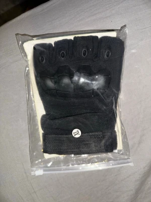 Svarta fingerlösa träningshandskar - Säljer ett par svarta fingerlösa handskar med förstärkta knogar och greppvänlig insida. Perfekta för träning eller gym. Justerbar kardborre vid handleden för bästa passform. Snygg och funktionell design för dig som vill ha extra skydd och stil. Storlek XL