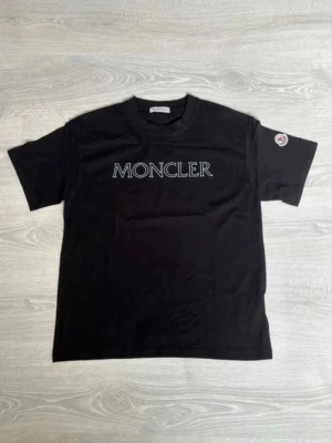 Svart Moncler t-shirt - Svart t-shirt från Moncler med stor vit logga på bröstet och Moncler-märke på ena ärmen. Klassisk rund halsringning och korta ärmar. Tillverkad i mjuk bomull för en skön känsla.