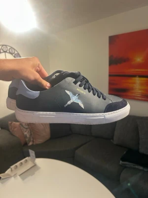 Mörkblå sneakers från Axel Arigato - Snygga mörkblå sneakers från Axel Arigato med vit sula och ljusblå detalj på hälen. Skorna har en broderad fågel på sidan och svarta skosnören. Materialet är en mix av läder och mocka för en stilren look.