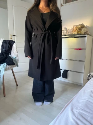 Svart kappa med knytskärp - Stilren svart kappa med knytskärp i midjan och bred krage. Kappan har en rak passform och långa ärmar, perfekt för kyligare dagar. Materialet ser ut att vara mjukt och något tjockare, vilket ger en snygg siluett och håller värmen. Köpt från missäy för 479kr. 