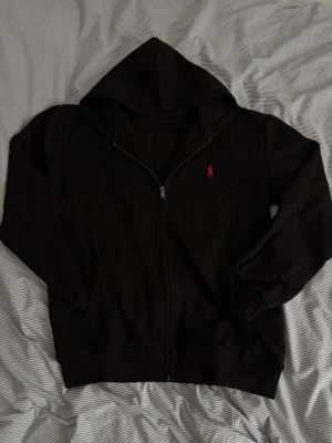 Svart hoodie från Polo Ralph Lauren - Svart hoodie med dragkedja från Polo Ralph Lauren. Storlek M, passar S/M. Tog ut snören för att tyckte att det passar inte, men man kan få dom med hoodien. Aldrig använd, sälja pga att den passar inte mig. 