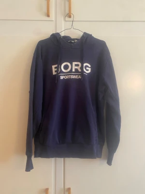 Mörkblå hoodie från  Björn BORG - Snygg mörkblå hoodie från Björn BORG med vit logga och texten 'SPORTSWEAR' på bröstet. Klassisk modell med huva och dragsnören samt magficka framtill. Perfekt för en sportig och avslappnad stil.