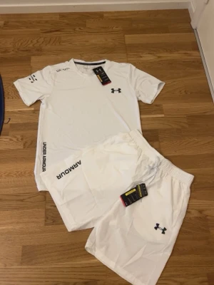 Vitt träningsset från Under Armour - Snyggt vitt träningsset från Under Armour med t-shirt och shorts. Båda plaggen har logga och tryck i svart. Tillverkade i lätt och snabbtorkande material som passar perfekt till gymmet eller löpning. Tidlös design med bekväm passform.