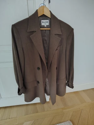 Brun oversized kavaj från NA-KD - Snygg brun oversized kavaj från NA-KD med klassisk krage, två knappar framtill och fickdetaljer. Kavajen har en rak passform och är helfodrad för extra komfort. Perfekt för en trendig och stilren look.