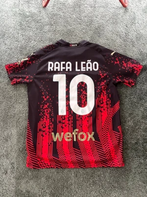 AC Milan matchtröja Rafa Leão #10 - Snygg AC Milan matchtröja med Rafa Leão och nummer 10 på ryggen. Tröjan är svart med röda digitala mönster och guldiga detaljer. Puma-logga, klubbmärke och sponsortryck på bröstet. Tillverkad i lätt och ventilerande material, perfekt för fotboll.