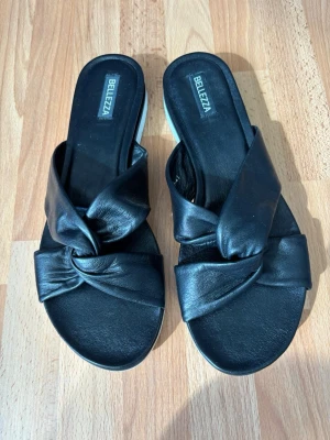 Svarta sandaler med knut från Bellezza - Stilrena svarta sandaler från Bellezza med breda remmar i konstläder som korsas och bildar en snygg knut framtill. Sandalerna har öppen tå och vit räfflad sula för extra grepp. Perfekta för en trendig och avslappnad look.