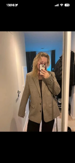Beige randig kavaj med bälte - Stilren beige kavaj med tunna vertikala ränder, knäppning framtill och två fickor. Kavajen har ett matchande bälte i midjan för en snygg siluett och klassisk krage. Perfekt för dig som gillar en tidlös och trendig look.