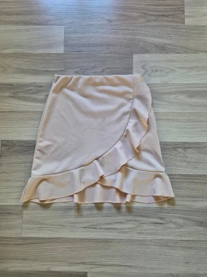 Beige omlottkjol med volang - Supersöt beige kortkjol med omlottdesign och volangkant. Kjolen har en mjuk och feminin look, perfekt för dig som gillar en romantisk stil. Resår i midjan för skön passform och lätt material som passar till många outfits.