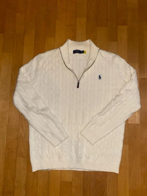 Vit kabelstickad tröja Polo Ralph Lauren - Kabelstickad vit tröja från Polo Ralph Lauren med halv dragkedja och blå broderad logga på bröstet. Tröjan har ribbade muddar och krage, perfekt för lager på lager. Klassisk och stilren design i mjukt material.