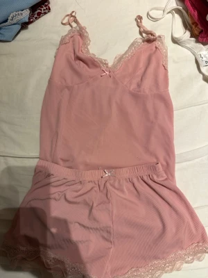 Rosa pyjamasset med spetsdetaljer - Sött rosa pyjamasset med linne och shorts. Linnet har tunna axelband, spetskant vid urringningen och en liten rosett framtill. Shortsen har elastisk midja, spetskant nedtill och matchande rosett. Mjukt, ribbat material som känns bekvämt mot huden.