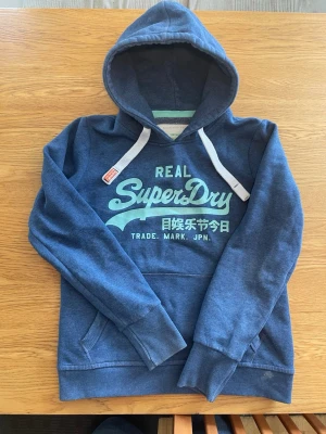 Blå Superdry hoodie med tryck - Str L men passar bättre S- M blå hoodie från Superdry med stort turkost tryck på bröstet som är glitter aktit. Har en liten fläck som syns på bilden längst ner fler bilder och mått kan skickas privat 😊 öppen för bud 
