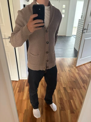 Beige stickad cardigan Less Worries - Säljer en beige stickad kofta med sjalkrage och stora knappar framtill. Koftan har två fickor och ribbade muddar vid ärmslut och nederkant. Perfekt för lager på lager och riktigt skön att ha på sig.