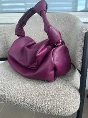 Lila handväska med knutdetaljer - Jättefin handväska från Gina Tricot i en mörk lila/burgundy nyans  Väskan har ett fint mjukt material, knutet handtag och en stilren, lite slouchy modell. Perfekt att bära till både vardag och fest. Skick: Väldigt fint använd skick. Det finns några små märken/slitage på undersidan av väskan, se bilder. De syns inte när väskan används och påverkar inte väskans funktion eller användning.