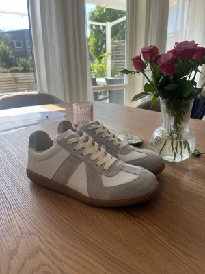 Vita och grå sneakers med mockadetaljer - Snygga sneakers i vitt och ljusgrått med beige skosnören och detaljer i mocka. Skorna har en klassisk låg siluett och en brun gummisula som ger en retrovibe. Perfekta för dig som gillar stilrena och tidlösa sneakers till vardagslooken.