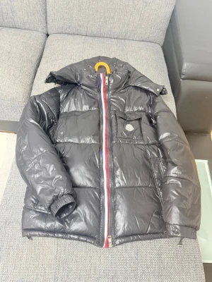 Svart dunjacka  - Snygg svart dunjacka från Moncler med glansig finish och klassisk Moncler-logga på bröstfickan. Jackan har en avtagbar huva, dragkedja med röd, vit och blå detalj samt elastiska muddar vid ärmsluten. Perfekt för kalla vinterdagar.