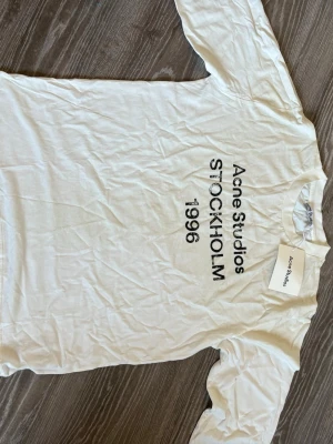 Vit Acne Studios t-shirt med tryck - Säljer en vit t-shirt från Acne Studios med svart texttryck 'Acne Studios STOCKHOLM 1996' på framsidan. Klassisk passform och rund hals. Perfekt till jeans eller shorts för en clean och stilren look.