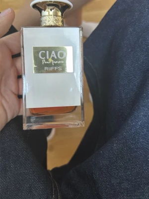 CIAO Pour Femme parfym från RiiFFS - CIAO Pour Femme parfym från RiiFFS i en elegant, rektangulär glasflaska med guldfärgad etikett och lock. Flaskan har en transparent design som visar den bärnstensfärgade vätskan inuti. Perfekt för dig som gillar lyxiga och stilrena dofter.