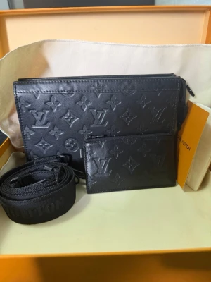 Svart Louis Vuitton handväska i läder - Svart handväska från Louis Vuitton i präglat läder med klassiskt monogrammönster. Väskan har en avtagbar axelrem och matchande liten plånbok. Perfekt accessoar för dig som gillar exklusiv och tidlös design.  (Kvitto finns)