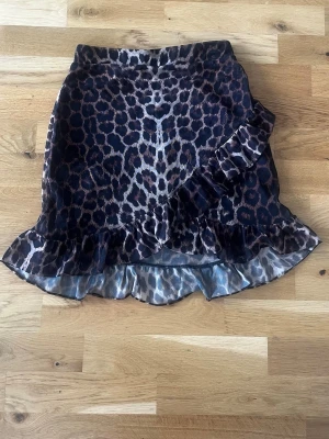 Leopardmönstrad volangkjol från NLY Trend - Säljer en kort kjol från NLY Trend med leopardmönster i bruna och svarta toner. Kjolen har volangdetaljer längs nederkanten och omlottkänsla framtill. Perfekt för dig som vill sticka ut med en trendig look.