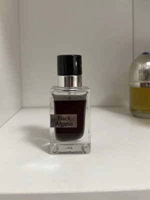 Black Afgano parfymflaska - Säljer en elegant parfymflaska med namnet Black Afgano. Flaskan är fyrkantig i genomskinligt glas med svart lock och mörk vätska inuti. Stilen är minimalistisk och modern, perfekt för dig som gillar exklusiva dofter.
