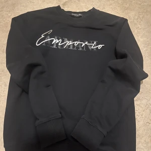 Svart sweatshirt från Emporio Armani - Svart sweatshirt från Emporio Armani med tryckt logga framtill i vitt och grått. Tröjan har rund halsringning, långa ärmar och ribbade muddar vid ärmslut och nederkant. Perfekt för en stilren och avslappnad look. Liten defekt på texten.