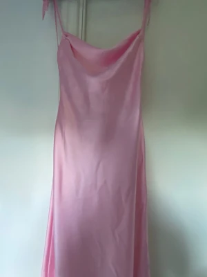Lång rosa satinklänning med smala band - Säljer en lång, ljusrosa klänning i satin med smala axelband och draperad urringning. Klänningen har en elegant, slät siluett och är perfekt för dig som vill ha en feminin och stilren look. Materialet ger ett snyggt fall och lätt glans.