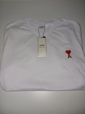 Vit t-shirt från Ami med rött hjärta - Säljer en stilren vit t-shirt från Ami med rund hals och ett broderat rött hjärta med bokstaven A på bröstet. T-shirten är i mjuk bomull och har en klassisk passform. Perfekt för dig som gillar minimalistisk design och snygga detaljer.