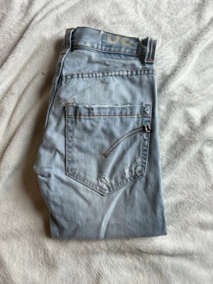 Dondupjeans - Säljer ett par ljusblå jeans med slitningar och råa detaljer på bakfickorna. Jeansen har klassisk femficksmodell, raka ben och kontrastsömmar. Perfekta för en avslappnad och trendig look.