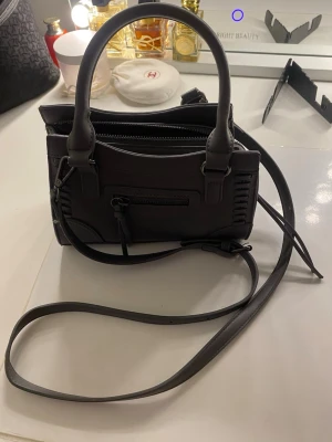 Mörkgrå handväska från Gina Tricot - Knappt använd, pris kan diskuteras. Snygg mörkgrå handväska från Gina Tricot i slätt konstläder. Väskan har både handtag och lång axelrem, samt en dragkedjeficka framtill och rymligt huvudfack med innerficka. Perfekt för dig som vill ha en stilren och praktisk accessoar. 