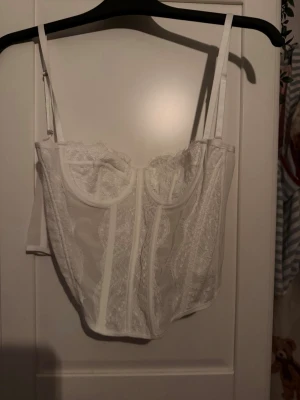 Vit spetskorsett med tunna axelband - Säljer en elegant vit korsett med tunna justerbara axelband och vackra spetsdetaljer. Korsetten har bygel under bysten och genomskinliga meshpartier som ger en feminin look. Perfekt för dig som vill ha något extra till din outfit.