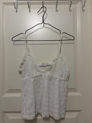 Vit spetslinnetopp med volang - Söt vit linnetopp med tunna axelband och volangdetalj längs ringningen. Toppen har ett broderat blommönster i spets och är perfekt för varma dagar. Lätt och luftig känsla, passar till både jeans och kjol.