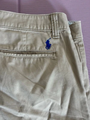 Beige chinos från Polo Ralph Lauren - Snygga beige chinos från Polo Ralph Lauren med klassisk passform. Byxorna har en diskret bakficka och det ikoniska blå broderade logotypen ovanför fickan. Tillverkade i mjukt bomullstyg som ger en skön känsla och stilren look.