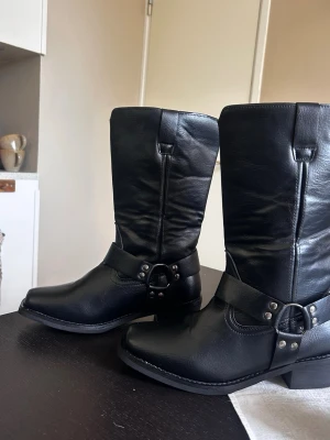 Svarta cowboy boots med spänne - Säljer ett par svarta cowboy boots i syntetmaterial med dekorativa spännen och nitar vid ankeln. Bootsens höga skaft och fyrkantiga tå. Skulle säga mellan storlek 40-41