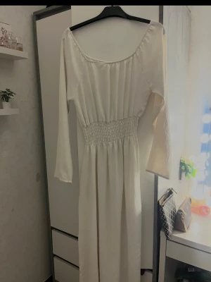 Vit långklänning med smock i midjan - Elegant vit långklänning med smockdetalj i midjan och långa ärmar. Klänningen har en vid, rundad ringning och ett mjukt, flytande fall. Perfekt för dig som gillar en enkel men stilren look.