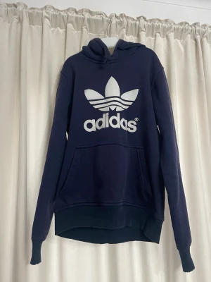 Mörkblå Adidas hoodie med logga - Snygg mörkblå hoodie från Adidas med stor vit Trefoil-logga på bröstet. Klassisk känguruficka framtill och huva. Tillverkad i mjukt material, perfekt för en avslappnad stil. Långa ärmar och ribbade muddar.