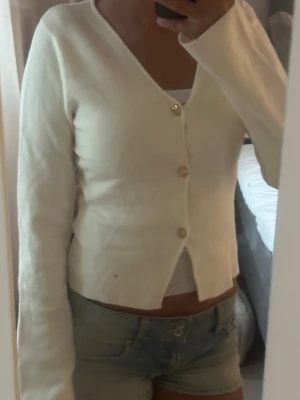 Vit kofta med knappar - Söt vit kofta med v-ringning och tre guldfärgade knappar framtill. Modellen är något croppad och har långa ärmar. Perfekt att slänga över ett linne för en enkel och stilren look. Storlek 158-164 men känns som en xs