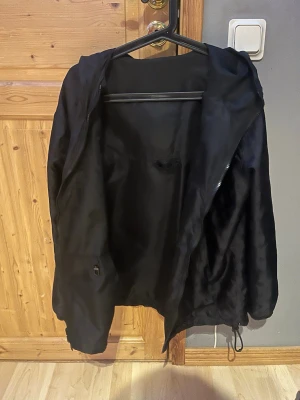 Svart vindjacka med monogrammönster - Snygg svart vindjacka med huva och dragkedja. Jackan har ett diskret monogrammönster över hela tyget och justerbar dragsko nedtill. Perfekt för dig som gillar stilrena detaljer och vill ha en lätt jacka till vardags.