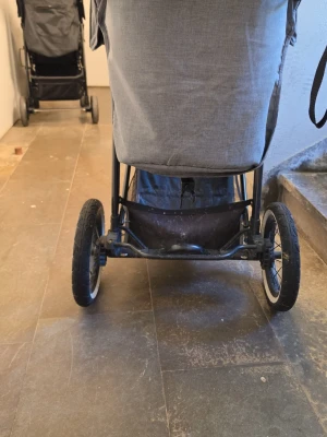 Grå barnvagn med stora hjul - Säljer en grå barnvagn med tre stora hjul och praktisk förvaringskorg undertill. Vagnen har mugghållare vid handtaget och ett robust chassi i svart metall. Perfekt för promenader och enkel att styra tack vare de stora hjulen.