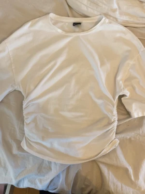 Vit ribbad topp med snörning - Snygg vit topp med ribbad struktur och snörning i sidan som ger en cool rynkad effekt. Kortärmad och med rund halsringning. Perfekt att styla till jeans eller kjol för en trendig look.Den är från gina tricot och är använd väldigt lite.