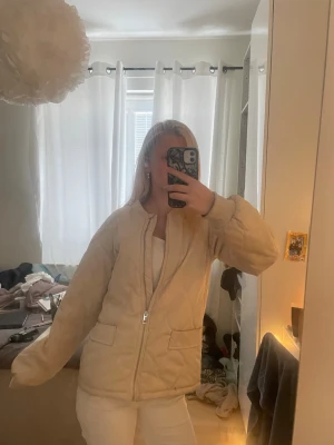 Beige pufferjacka med dragkedja - Säljer en beige pufferjacka med rak passform och två stora fickor framtill. Jackan har en enkel design utan synligt märke, elastiska muddar vid ärmsluten och stängs med dragkedja. Perfekt för kalla dagar och ger en clean look.