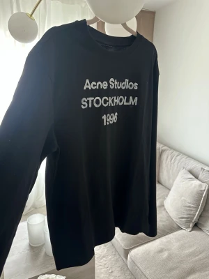 Svart långärmad topp Acne Studios - Svart långärmad topp Acne Studios med trycket 'Acne Studios STOCKHOLM 1996' i vitt framtill. Klassisk rund halsringning och relaxed fit. Perfekt för dig som gillar stilrena och trendiga plagg.