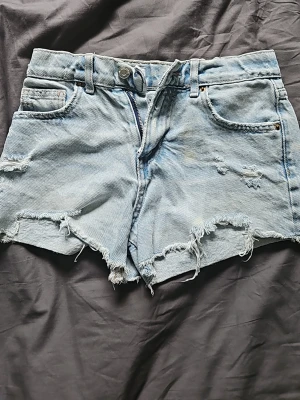 Ljusblå slitna jeansshorts - Säljer ett par ljusblå jeansshorts med råa kanter och slitna detaljer. Shortsen har klassisk femficksdesign, bälteshällor och knappgylf. Perfekta för sommaren och ger en avslappnad vibe.