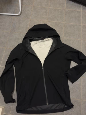 Svart vindjacka från uniqlo - Snygg svart vindjacka med hel dragkedja och huva. Jackan har en minimalistisk design och är tillverkad i ett tunt, vindtätt material med ljus insida. Perfekt för blåsiga dagar och enkel att matcha med olika outfits.