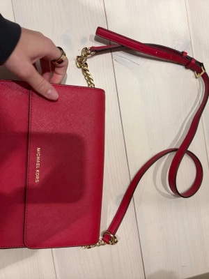 Röd axelväska från Michael Kors - Snygg röd axelväska från Michael Kors i slätt läder med guldfärgade detaljer och kedja på axelremmen. Väskan har flera fack inuti och justerbar rem. Perfekt för att lyfta vilken outfit som helst.