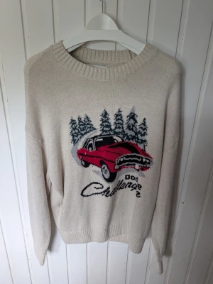 Beige stickad tröja med bilmotiv - Stickad tröja från Hollister i boxy fit, beige färg med ett stort motiv av en röd bil och granar samt texten 'Challenger' framtill. Tröjan har rund halsringning och ribbade muddar. Perfekt för dig som gillar unika prints och retrovibbar.