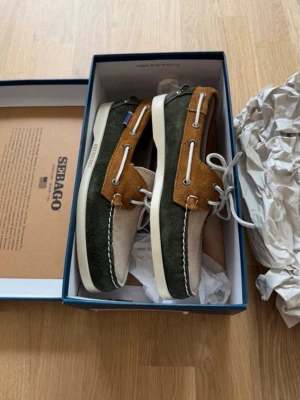 Sebago seglarskor i mocka - Säljer ett par Sebago seglarskor i mocka med snygg färgkombination av mörkgrönt, beige och brunt. Skorna har vita snören och detaljer samt en vit sula. Klassisk design med robust känsla och Sebago-logga på sidan.