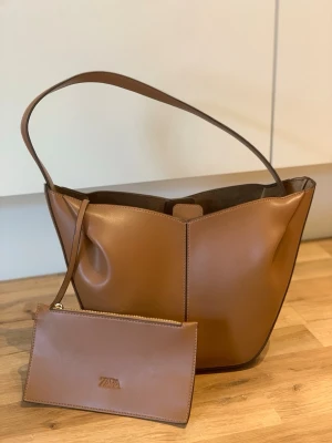 Brun bucket bag från Zara - Snygg brun bucket bag från Zara i slätt konstläder. Väskan har en enkel axelrem och kommer med en matchande liten clutch med dragkedja. Perfekt för dig som gillar stilrena och praktiska accessoarer. Rymmer allt du behöver till vardags.