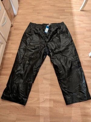 Svarta vida adidas byxor.  - Säljer ett par svarta mjukisbyxor med bred passform och resår i midjan. Byxorna är gjorda i ett glansigt material som ger en sportig vibe. Perfekta för chill dagar eller när du vill ha något riktigt bekvämt. Storlek XXXL. 